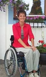 Lena Käld sitter i en rullstol. I bakgrunden syns en husvägg och hon ler mot kameran.
